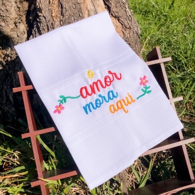 Toalha branca bordada com a frase 'amor mora aqui' e flores, sobre estrutura de madeira junto a um tronco de árvore