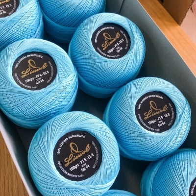 Oito novelos de fio de algodão azul claro em bobinas redondas com etiquetas pretas numa caixa cinza