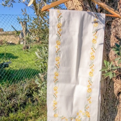 Toalha branca com bordados amarelos e verdes pendurada num cabide numa árvore