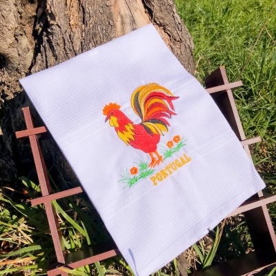 Toalha branca bordada com galo colorido e texto PORTUGAL sobre estrutura de ferro ao lado de árvore