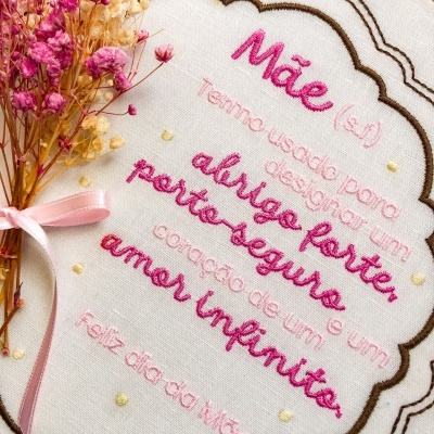 Bordado em tecido branco com texto em rosa e vermelho e ramo de flores secas com fita rosa.