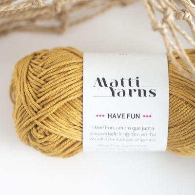 Bola de fio amarelo mostarda com etiqueta 'Matti Yarns HAVE FUN' junto a rede bege