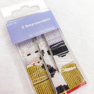 Embalagem de agulhas Smyrnanadeln de metal com cartão azul e padrão listrado colorido.