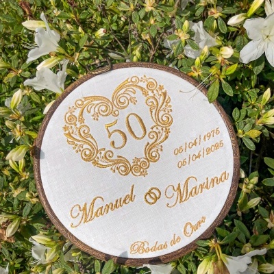 Bordado redondo branco com design dourado de coração e texto comemorativo rodeado de folhagem e flores brancas