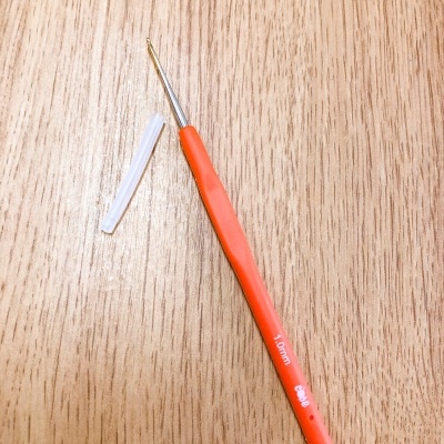 Agulha de crochet laranja com ponta metálica dourada e protetor branco sobre madeira