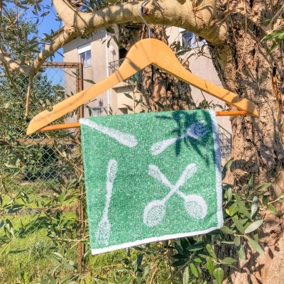 Toalha verde com desenhos brancos de utensílios de cozinha pendurada num cabide de madeira ao ar livre