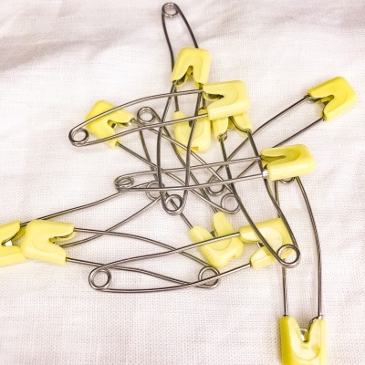 Vários alfinetes de segurança metálicos com ponta amarela sobre pano branco.