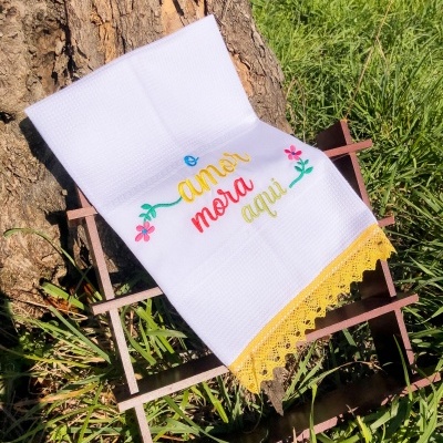Toalha branca bordada com frase colorida e renda amarela sobre estrutura de madeira na natureza