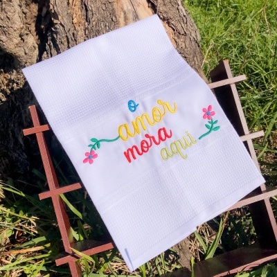 Toalha branca com bordado colorido 'o amor mora aqui' apoiada em estrutura de madeira frente a tronco de árvore