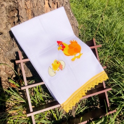 Toalha branca com bordado de galinha e pintainho, com renda amarela, sobre estrutura metálica, em ambiente ao ar livre.