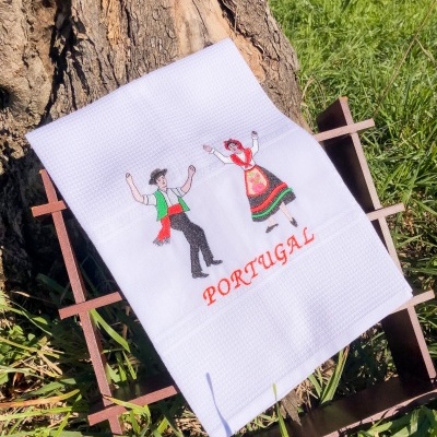 Toalha branca bordada com casal tradicional português e texto PORTUGAL sobre suporte de madeira junto a árvore.