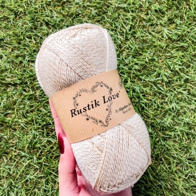 Skein de fio bege claro com rótulo Rustik Love em mão sobre relva verde
