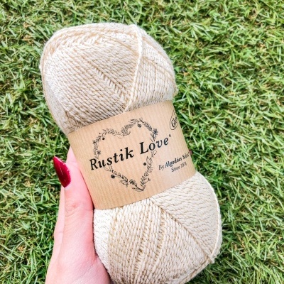 Novelo de lã bege com rótulo Rustik Love, segurado por mão sobre relva verde
