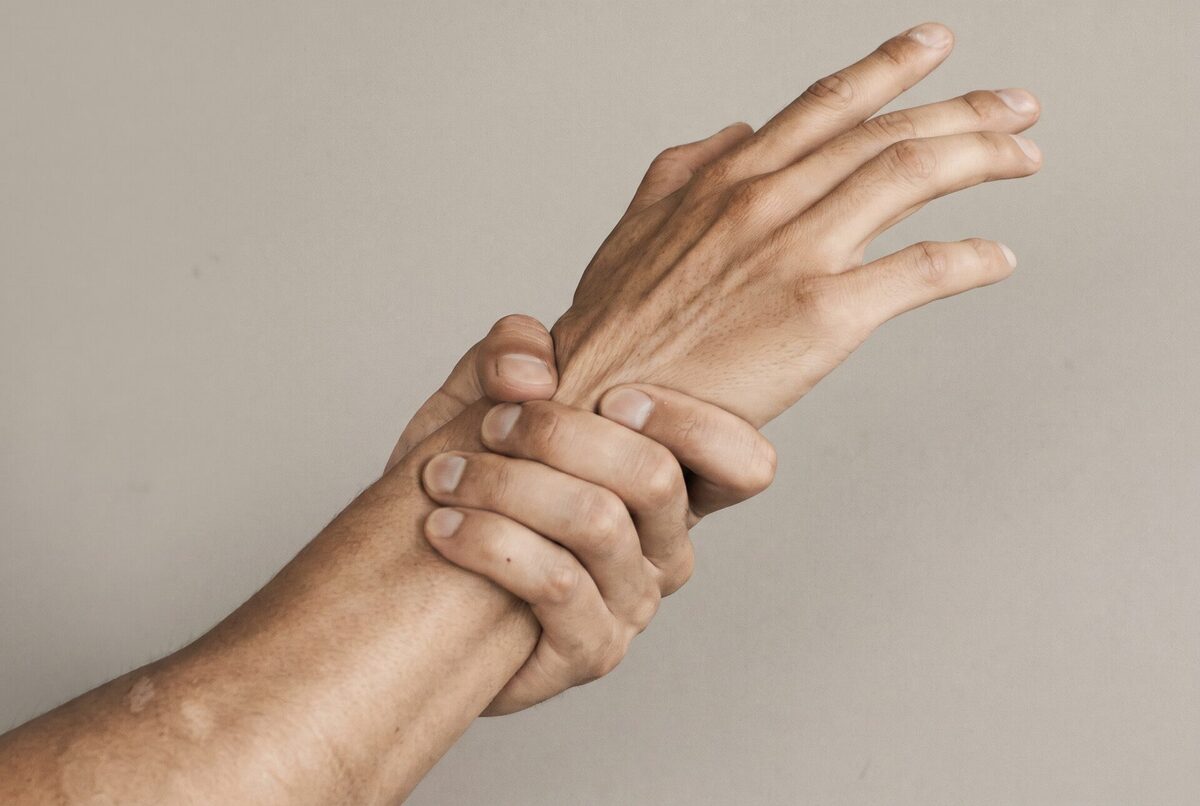 Duas mãos humanas, uma segurando o pulso da outra, fundo bege neutro.