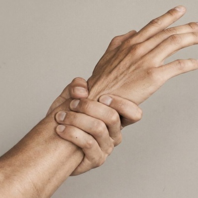 Duas mãos humanas, uma segurando o pulso da outra, fundo bege neutro.