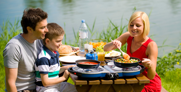 Família sentada à mesa de madeira ao ar livre junto a um lago com comida