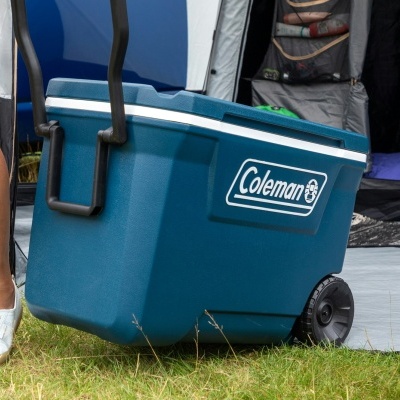 Caixa térmica azul Coleman com pega e rodas pretas sobre relva junto a tenda