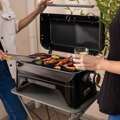 Churrasqueira portátil preta a grelhar legumes e carne em mesa dobrável