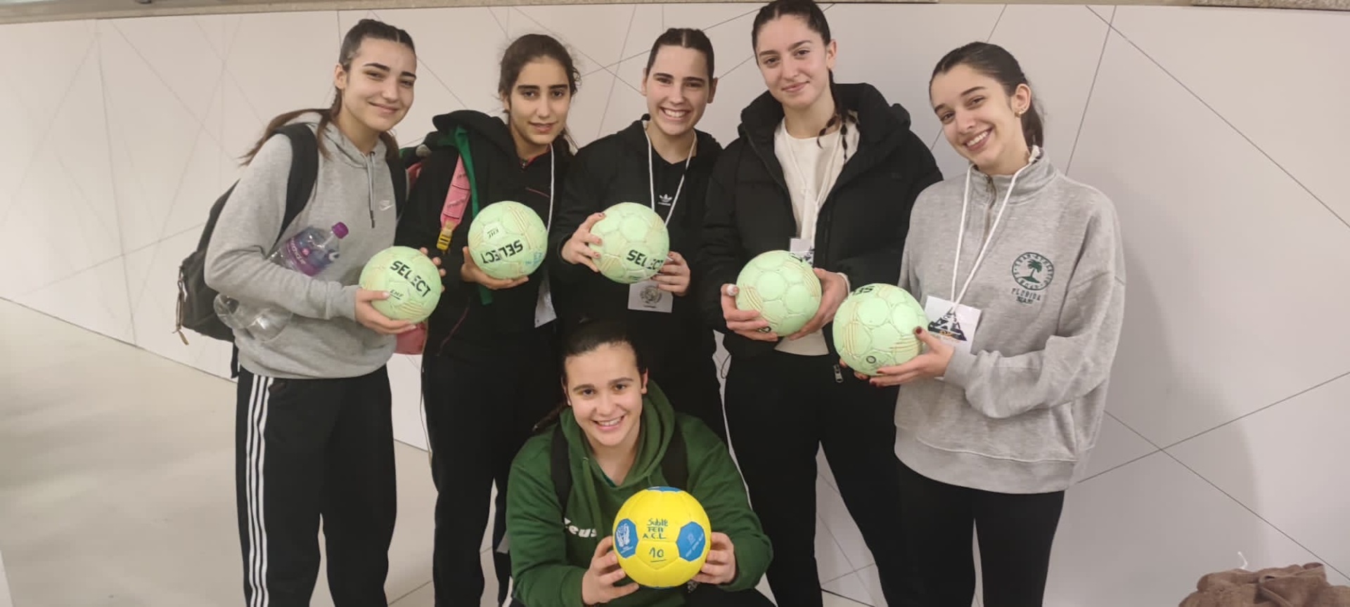 Grupo de seis mulheres com roupas desportivas segurando bolas de andebol verdes e uma amarela num ambiente interior branco