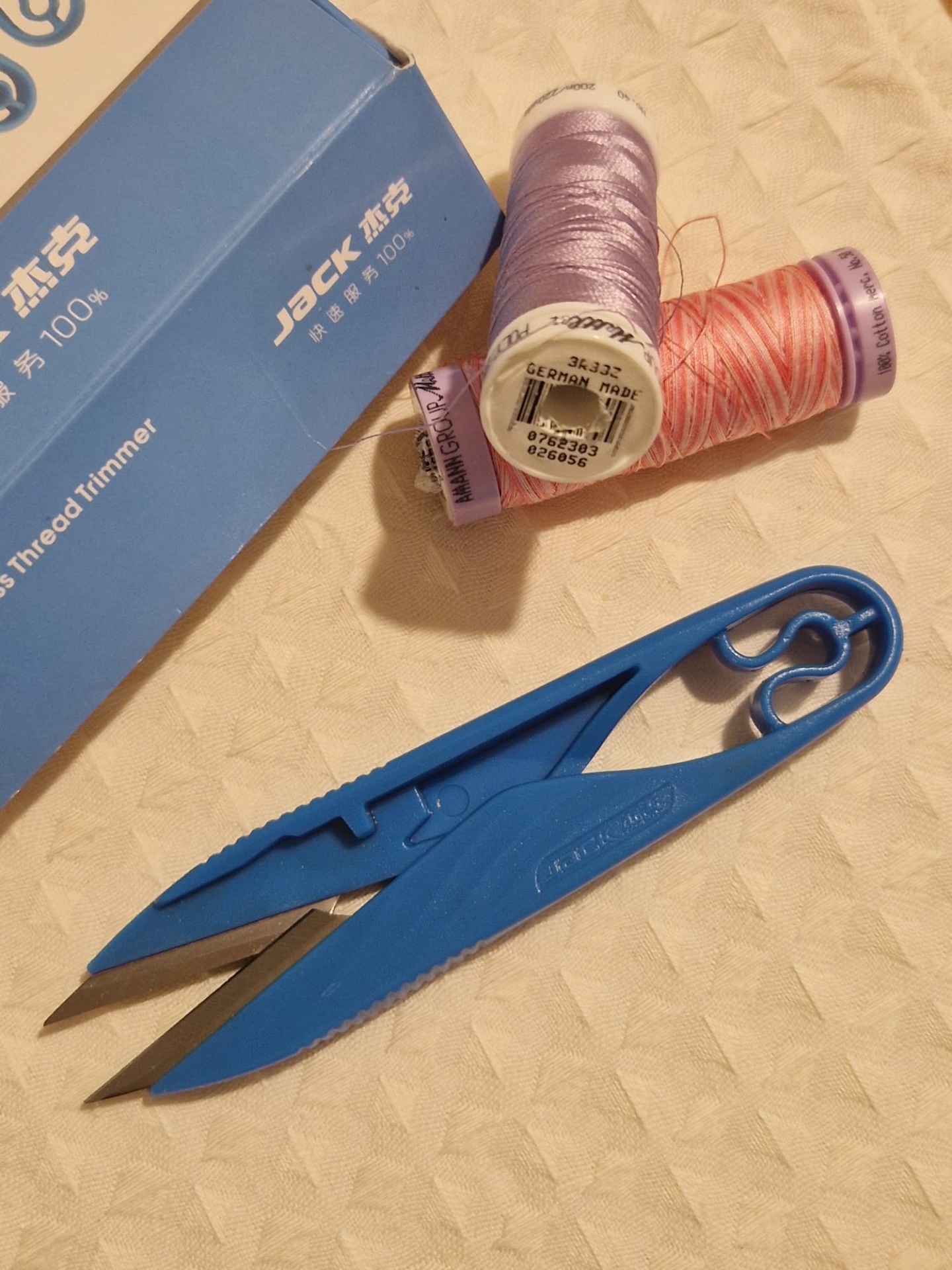 Tesoura corta pontas JACK Tesoura azul, bobinas de linha e caixa azul com texto em tecido bege.