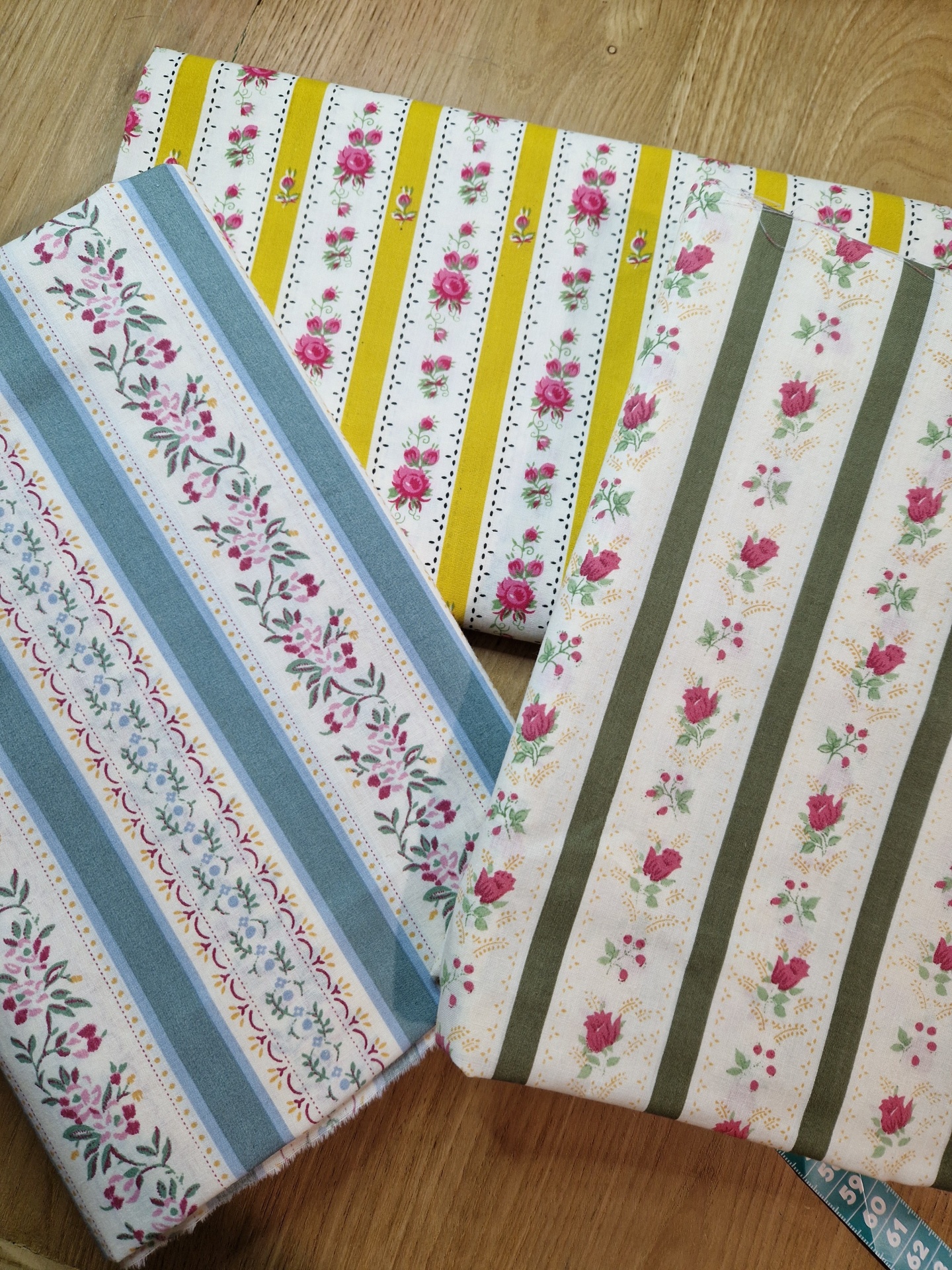 Tecido Chita Verde Azeitona Três tecidos florais com padrões de riscas em amarelo, azul e verde sobre fundo branco, sobre madeira.