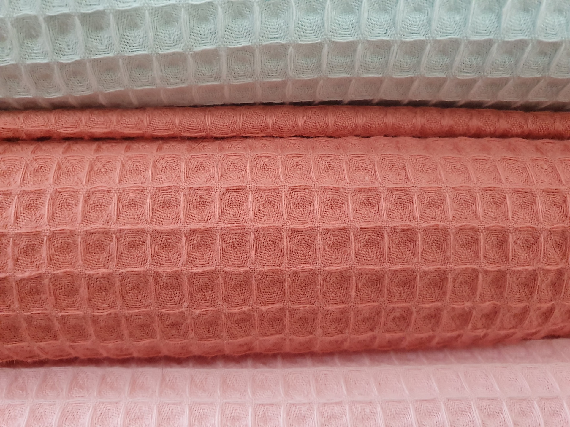 Tecido Waffle Coral Escuro Três tecidos dobrados com padrão de grade em relevo nas cores branco, vermelho tijolo e rosa claro.