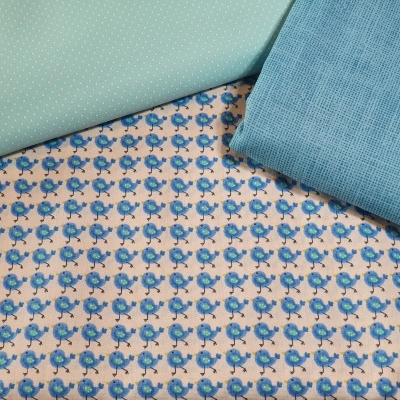 Três tecidos diferentes: verde claro com pontos, azul claro texturizado e padrão de pequenos pássaros azuis sobre fundo bege.