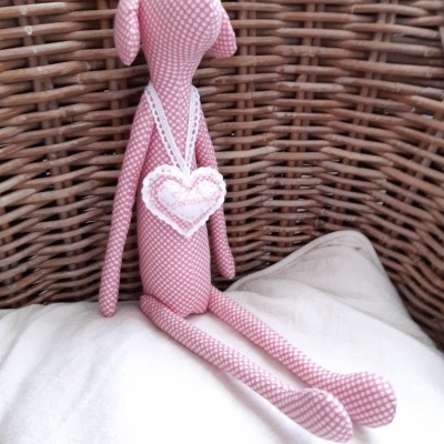 Boneco de tecido rosa com coração branco pendurado no pescoço sentado em almofada