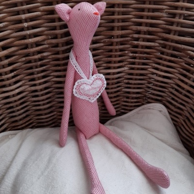 Boneco de tecido rosa com padrão xadrez sentado num cesto de vime com coração pendurado no pescoço