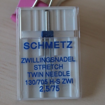 Agulha dupla Schmetz para costura em caixa transparente com texto azul, em superfície de madeira clara.
