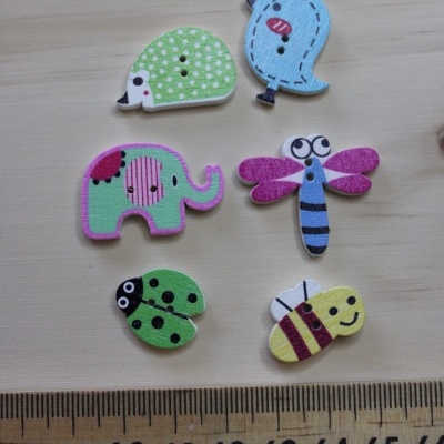 Seis botões decorativos em madeira com desenhos de animais e insectos coloridos em cima de uma superfície de madeira clara junto a uma régua.