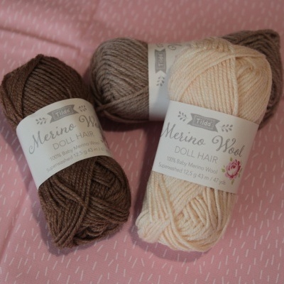 Três novelos de lã Merino Wool Doll Hair em várias cores sobre fundo rosa com padrão de riscos brancos.