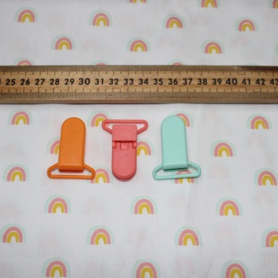 Três clips de chupeta coloridos em pano branco com padrão de arcos-íris e régua de madeira