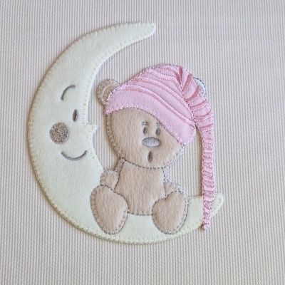 Aplicação bordada de uma lua branca e um urso castanho com gorro rosa sobre tecido bege