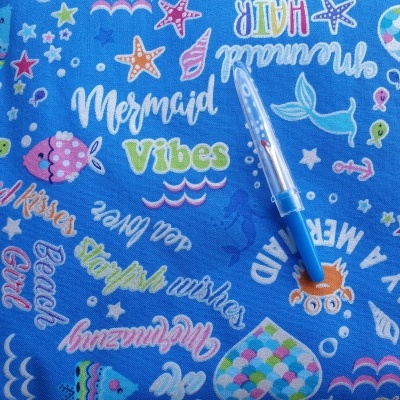 Tecido azul com padrão colorido de sereias e mar, com caneta azul sobre ele