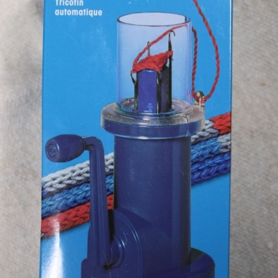 Embalagem azul mostrando máquina de tricô azul com manivela e fios trançados vermelhos