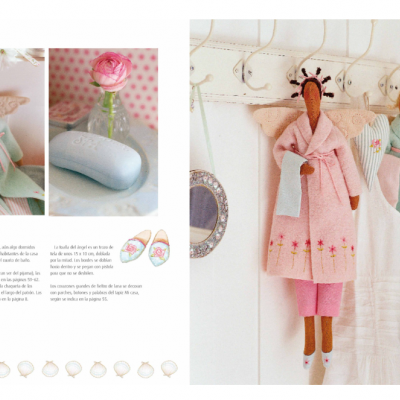 Anjos de pano em bata azul e rosa pendurados em ganchos, sabonete azul e flor rosa em vaso, página de livro com texto em português