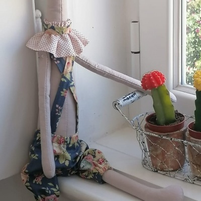 Boneco de giraffe de tecido com roupas floridas sentado na janela ao lado de cactos.