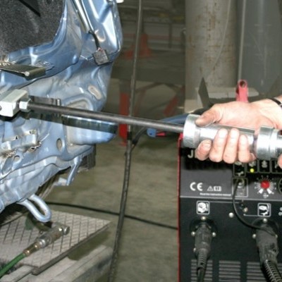 Ferramenta elétrica com empunhadura cinza e haste metálica usada numa estrutura azul com máquina preta ao fundo