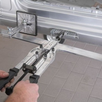 Ferramenta metálica com alças pretas presa à carroçaria de um carro prateado num chão de ladrilhos cinzentos