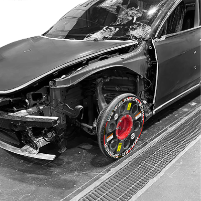 Carro cinza escuro com a frente danificada e roda dianteira com dispositivo colorido em ambiente industrial.