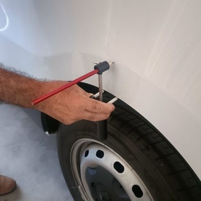 Pessoa a usar ferramenta vermelha e preta numa viseira branca de veículo com pneu e jante metálica num chão cinzento.