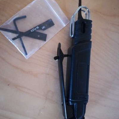 Dispositivo negro portátil com gancho metálico e ferramentas num saco plástico sobre madeira