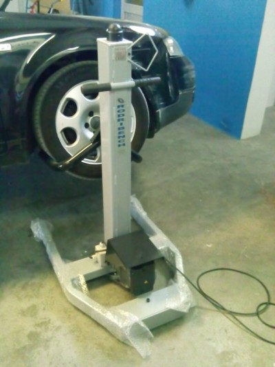 Aparelho metálico cinzento com cabo elétrico junto a uma viatura preta numa garagem.