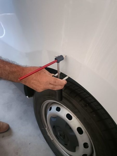 Pessoa a usar ferramenta vermelha e preta numa viseira branca de veículo com pneu e jante metálica num chão cinzento.