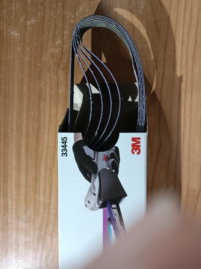 Correias abrasivas pretas 3M 33445 em embalagem branca sobre mesa de madeira.