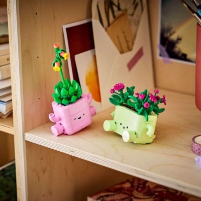 Dois vasos de flores decorativos em forma de cubos sorridentes, cor-de-rosa e verde, com flores, numa prateleira de madeira