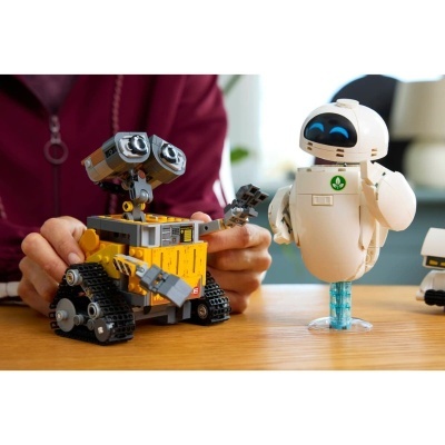 Dois robots de brinquedo tipo Lego sobre mesa de madeira, um amarelo e cinza com lagartas, outro branco com olhos azuis, mão de pessoa a interagir