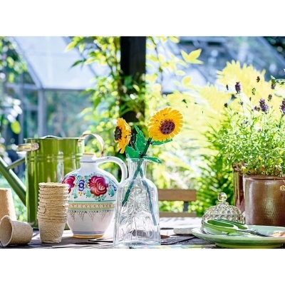 Vaso com girassóis de plástico, jarra cerâmica decorativa, regador verde, planta em vaso de barro e pratos verdes numa mesa de jardim