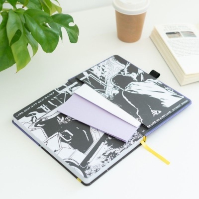 Caderno aberto com ilustrações em preto e branco e folhas soltas em mesa com planta, livro e copo de papel.